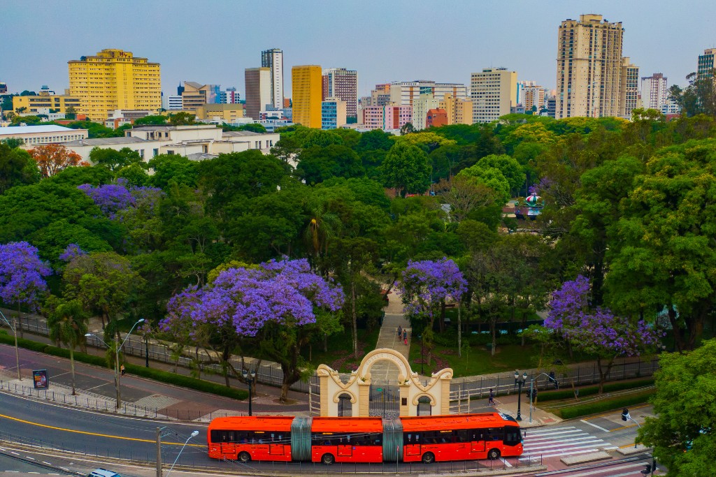 Vista aérea do Passeio Público em Curitiba, com jacarandás em flor, arcos de entrada e horizonte urbano